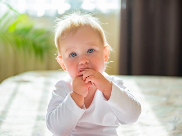 A baby is sitting and looking forward with their hands covering their mouth