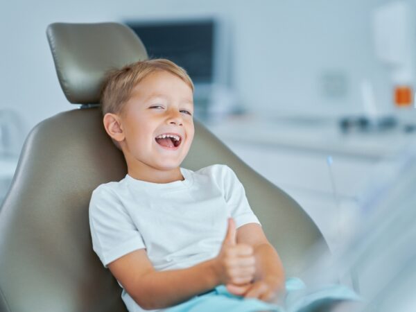 A smiling boy is sitting in a chair at the orthodontist doing a thumbs up